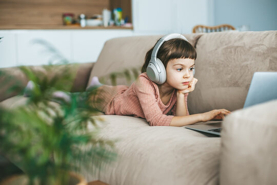 With headphones over her ears, a little girl lying on a beige sofa, engrossed in the captivating scenes unfolding on her laptop screen