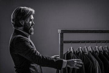 Male beauty and fashion. Bearded man tailoring clothes. Male suits hanging in a row. Men clothing, boutiques. Tailor, tailoring. Stylish men's suit. Man suit, tailor in his workshop. Black and white