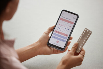 Over shoulder closeup of young woman holding smartphone with calendar app on screen and pack of birth control pills - 787130516