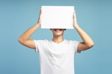 Smiling positive boy with braces holding white blank screen, covering face, on blue background