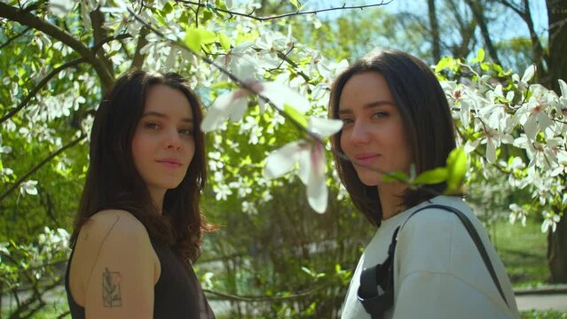 Beautiful women looking smiling at camera. Couple of female friends in sunlight surrounded by soft haze of magnolia blossom petals in garden. Cinematic slow motion shot