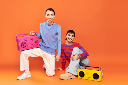 two cheerful good looking gay men with vibrant makeup posing with tape recorders on orange backdrop