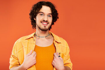 merry appealing gay man with dark hair and vibrant makeup posing on orange backdrop, pride month