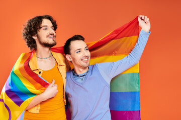 cheerful appealing young gay men in cozy attires with rainbow flag on orange backdrop, pride month