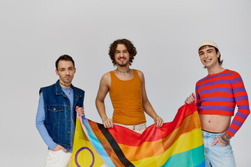 three joyous handsome gay men in vibrant clothes posing with rainbow flag and looking at camera