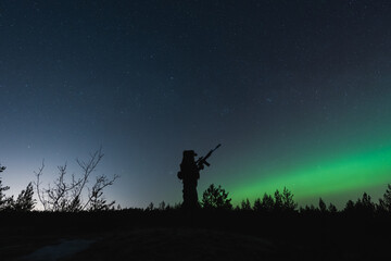 Soldier with a night vision device and a rifle with a suppressor in the forest against the background of the starry sky and northern lights.