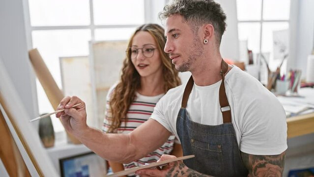 A woman and a tattooed man in aprons working together with paintbrushes in a bright art studio.