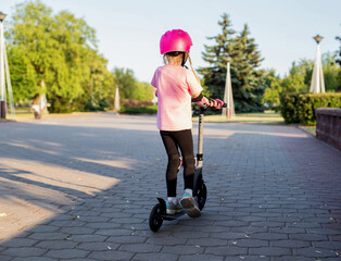 A girl 7 years old in a helmet and a pink injury protection kit rides a scooter in the park, sunny day. Active sports and recreation. Safety when riding bicycles and scooters. Copy space for text - 787161340
