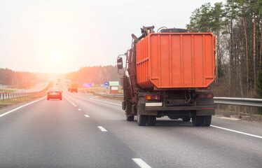 An orange garbage truck drives along an asphalt country road in the spring against the backdrop of sunset. Garbage collection concept, copy space for text