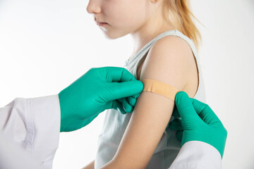 Doctor's hands in green medical gloves stick a plaster on the shoulder of a girl child after vaccination against hemophilus influenzae and pneumococcal. Copy space for text, revaccination - 787161540