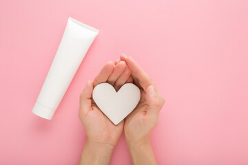 Young adult woman hands holding white heart shape. Cream tube on light pink table background. Pastel color. Care about clean and soft female body skin. Closeup. Point of view shot. Top down view. - 787164582