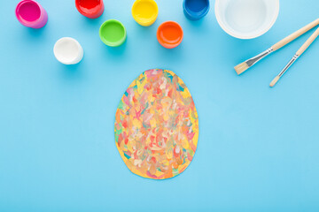 Painted colorful egg on paper, paintbrush and gouache bottles on light blue table background. Pastel color. Closeup. Making easter decoration elements. Top down view. - 787164587
