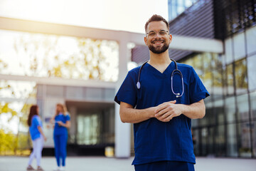 A male nurse is standing outdoors wearing scrubs and a stethoscope. He is smiling and looking at...