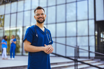 Portrait of smiling male nurse standing in hospital. Portrait Of Male Doctor Wearing Scrubs Standing In Modern Hospital Building. Male healthcare worker smiling outside, portrait - 787172348