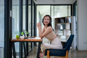 Smiling young woman with headphones sitting at a table with a laptop, enjoying music during study time at home.