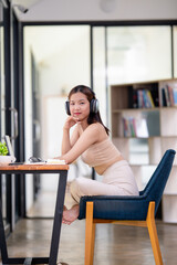 Smiling young woman with headphones sitting at a table with a laptop, enjoying music during study time at home.