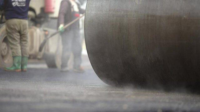 Steamroller compactor cylinder flatten out the new asphalt with steam evaporationg in slow motion. Industrial construction concept
