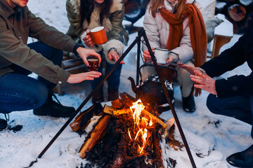 Group of friends gathering around bonfire in backyard, drinking tea and warming hands