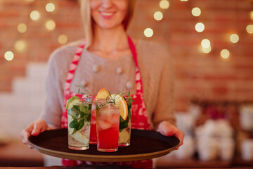 Smiling waitress in colored apron holding tray with alcoholic drinks and various cocktails