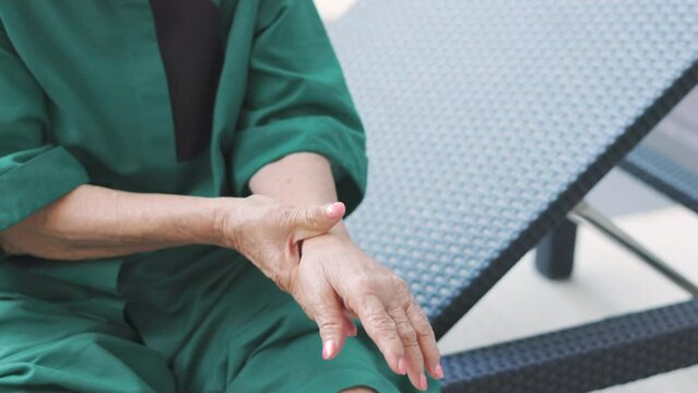 Close up 4K of hands holding painful wrist and arms of senior woman who is sitting on chair outdoor shows concept of health care problem of elderly people which needs social support for joint surgery.