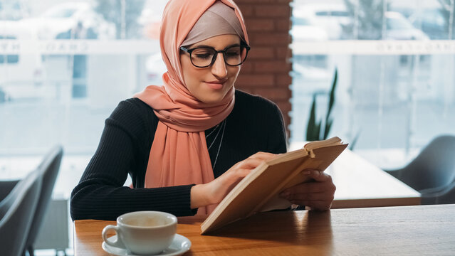 Cafe book. Reading leisure. Concentrated woman in hijab and glasses university student studying learning novel at cozy coffee shop free space.