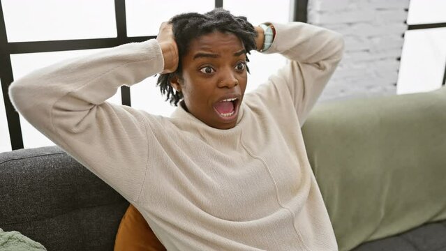 Shocked young black woman with dreadlocks goes bonkers at home, sitting on the sofa! hands on head, open mouthed in surprise, scared out of her wits!