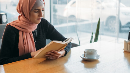 Cafe reading. Book leisure. Serene cheerful woman in hijab enjoying novel story weekend relaxing at...