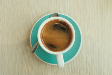 Cup of coffee on a textured wooden table. The coffee is in a white cup on a teal colored saucer....