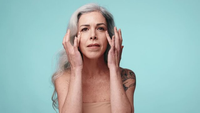 Senior lady showing under-eye skincare routine against a blue background. Concept of beauty and aging.