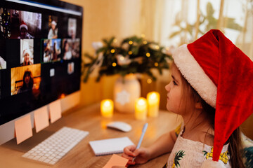 Little cute girl in santa hat sits at table, looks at monitor screen and writes or draws on paper...