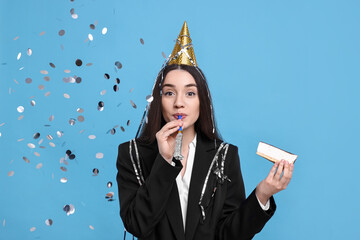 Woman in party hat with blower, piece of tasty cake and flying confetti on light blue background