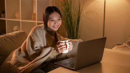 Hygge evening. Home rest. Weekend leisure. Relaxed happy woman watching movie on laptop with hot tea alone on cozy couch.