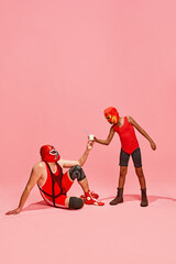 Boy in superhero dressed red costume gave glass of milk to his older, bigger partner against pink studio background. Concept of pop art, generation difference, costume festivals, competitions. Ad