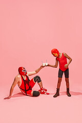Masked wrestler in red gear seated, toasting milkshakes with standing partner against pink studio background. Concept of pop art, generation difference, costume festivals, competitions. Ad