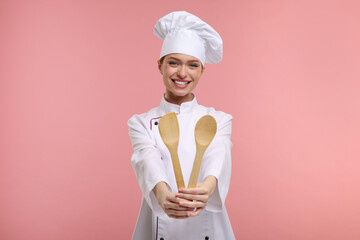 Happy chef in uniform holding wooden spatula and spoon on pink background