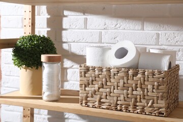 Toilet paper rolls in wicker basket, floral decor and cotton pads on wooden shelf near white brick wall
