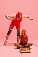 Wrestler in red performs pose above a seated partner with mask, who eyeing a pizza. against pink studio background. Concept of pop art, generation difference, costume festivals, competitions. Ad