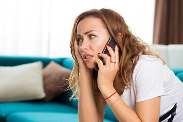 Portrait of an unhappy young woman talking on cellphone in her apartment  - 787214910