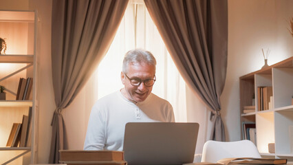 Online communication. Mature man. Digital life. Happy senior male working laptop on desk light home...