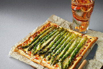 Opened pie with asparagus and cheese next to glass of lemonade with ice on gray background