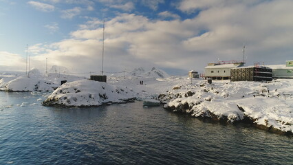 Close up view, low above water drone flight near Antarctic polar station - Vernadsky Base. People on ocean water coast landscape. Snow covered settlement. Slow aerial flight