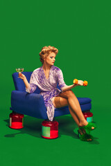 Girl drinks cocktail while sitting on vintage chair that has jars of vitamins instead of legs against vibrant green background. Concept of sport, competition, active lifestyle, hobby, recreation. Ad