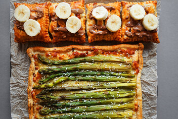 Opened asparagus and cheese pie next to banana puffs on parchment paper on gray background