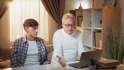 Diverse generation. Computer problem. Family quarrel. Skeptic son looking to shocked shrugging shoulders father using laptop home interior. - 787224951