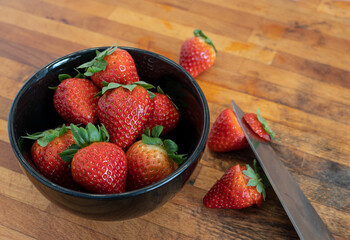 Strawerries preparation in the kitchen. Black bowl full of strawberries and a knife cutting the...