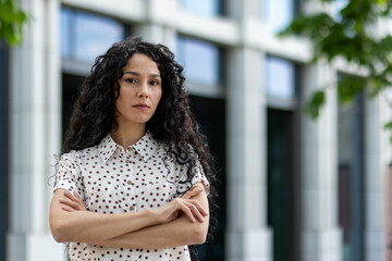 Portrait of a determined Hispanic woman, standing confidently with her arms crossed. The background features modern urban architecture, emphasizing a professional atmosphere.