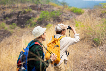 active asian senior couple hiking together on summer time,a female looking into binoculars and smiling,concept of elderly pensioner lifestyle,family relationship,activity,travel,adventure in nature