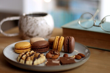 Plate with macarons, cookies, chocolate and nuts, cup of tea or coffee, book and reading glasses on...