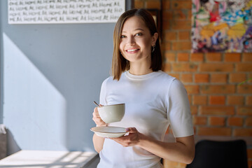 Young beautiful woman holding cup of coffee, looking out window, gray wall copy space