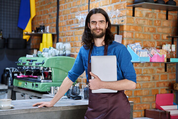 Young man in apron, food service worker, small business owner holding laptop